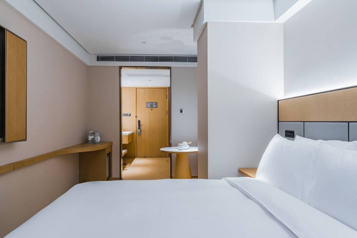 A hotel room with a bed, a bedside table, a small round table with a cup, a mirror, and a door leading to a bathroom.