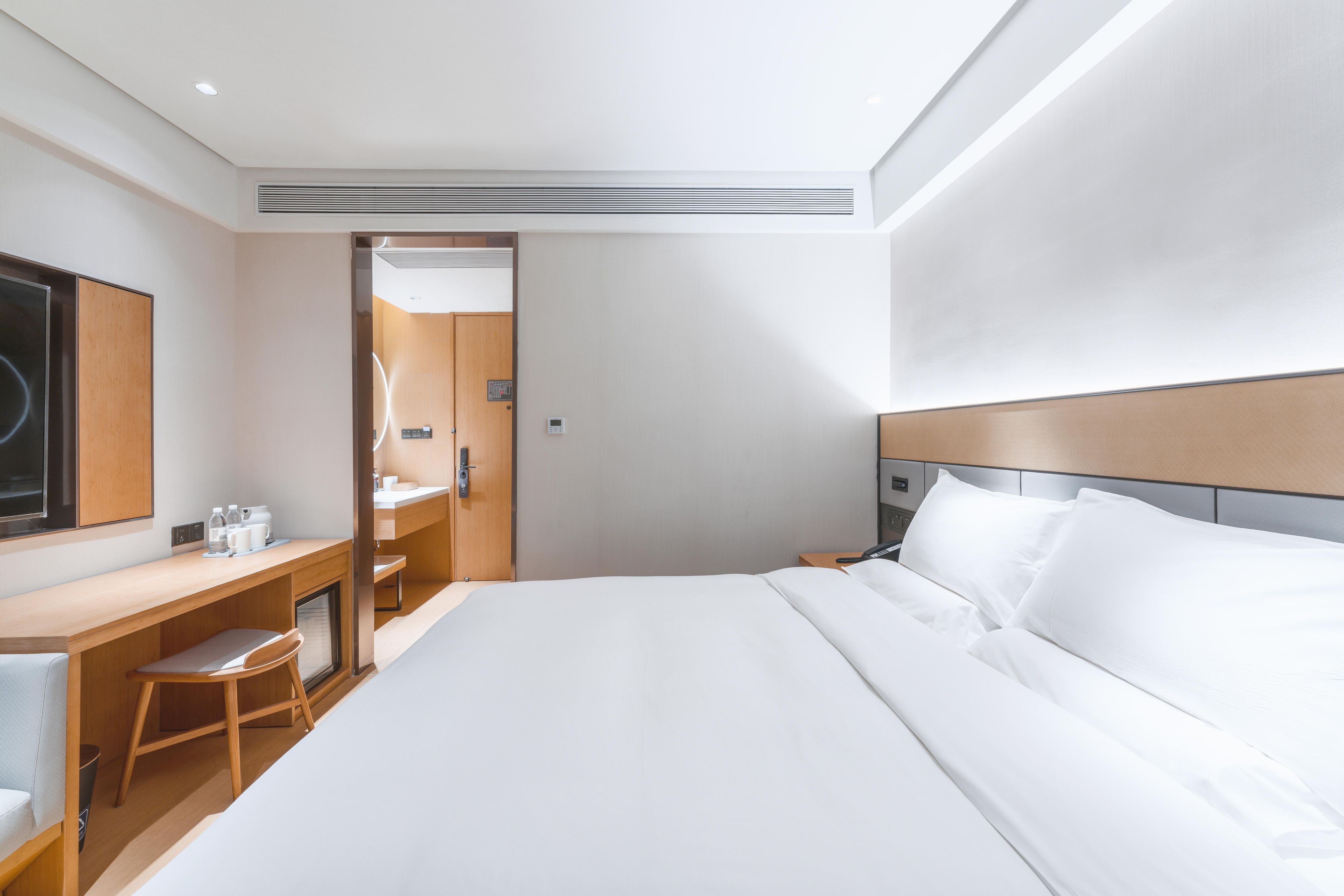 A modern hotel room with a large bed, a desk with a chair, a mirror, and a bedside table.