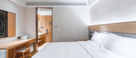 A modern hotel room with a large bed, a desk with a chair, a mirror, and a bedside table.