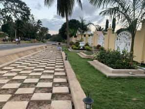 A paved pathway leading to a residential area with a well-maintained lawn, decorative plants, and a white gate.