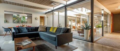 A modern living room with a sectional sofa, a coffee table, and a dining area visible through glass doors.