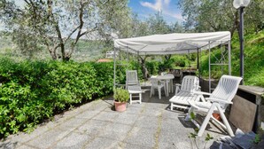 Un patio exterior con sillas y una mesa blanca bajo un toldo, rodeado de follaje y árboles.