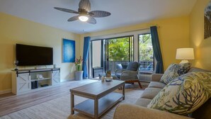 A living room with a flat-screen TV, a ceiling fan, a sofa, a coffee table, and a view of greenery outside. - LOW August   rates  A/C-Free Parking-No Resort Fees (Kihei)