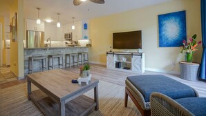 A modern living room with a wooden coffee table, a blue sofa, a TV, and a kitchen area with a bar. - LOW August   rates  A/C-Free Parking-No Resort Fees (Kihei)