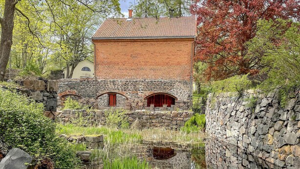 Ein traditionelles Backsteingebäude mit einem Ziegeldach, einer Steinmauer und einem kleinen Teich im Vordergrund.