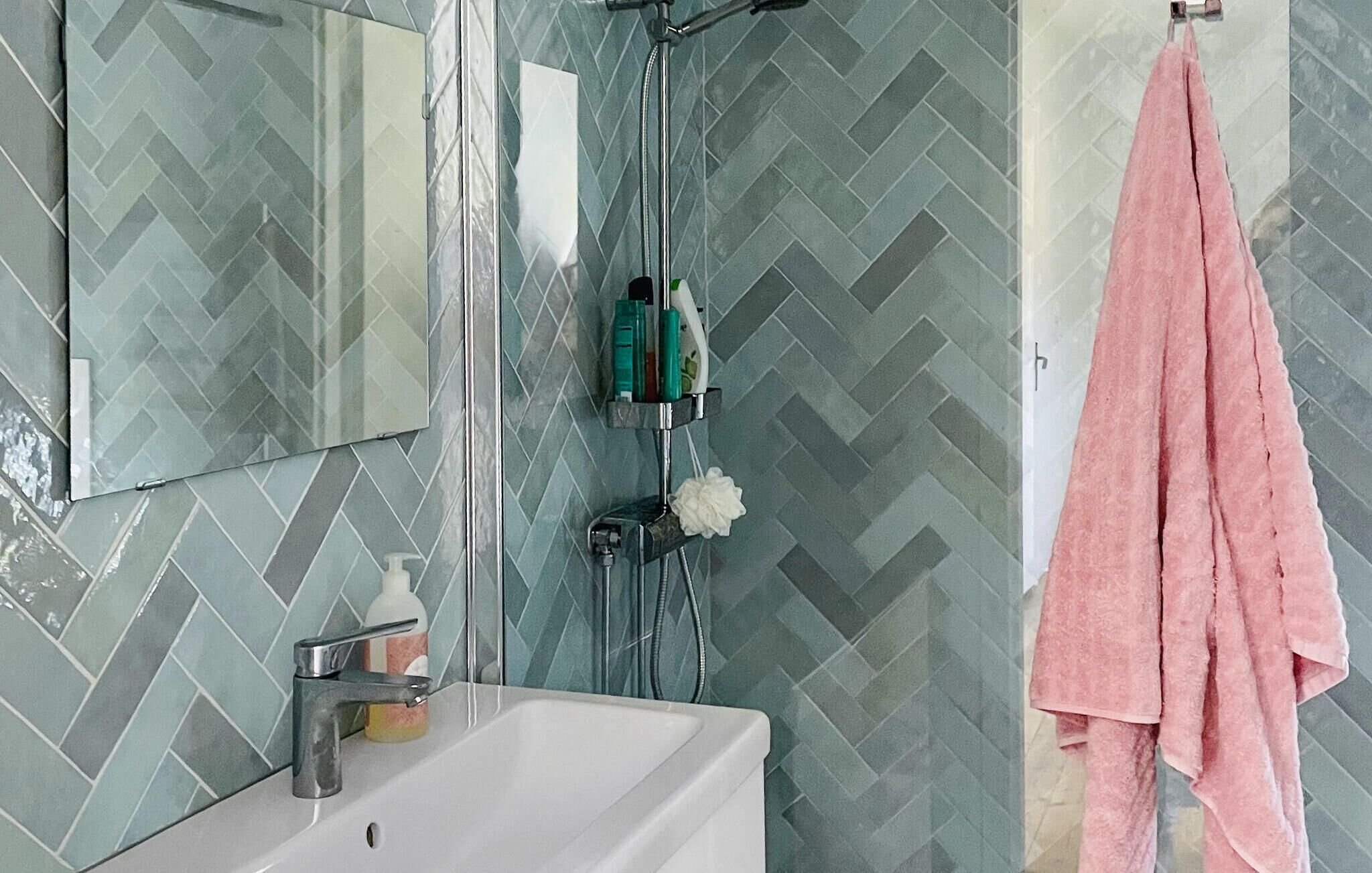 A bathroom with a white sink, a mirror, and a shower with a glass door. There are toiletries on the sink and a pink towel hanging on the wall.
