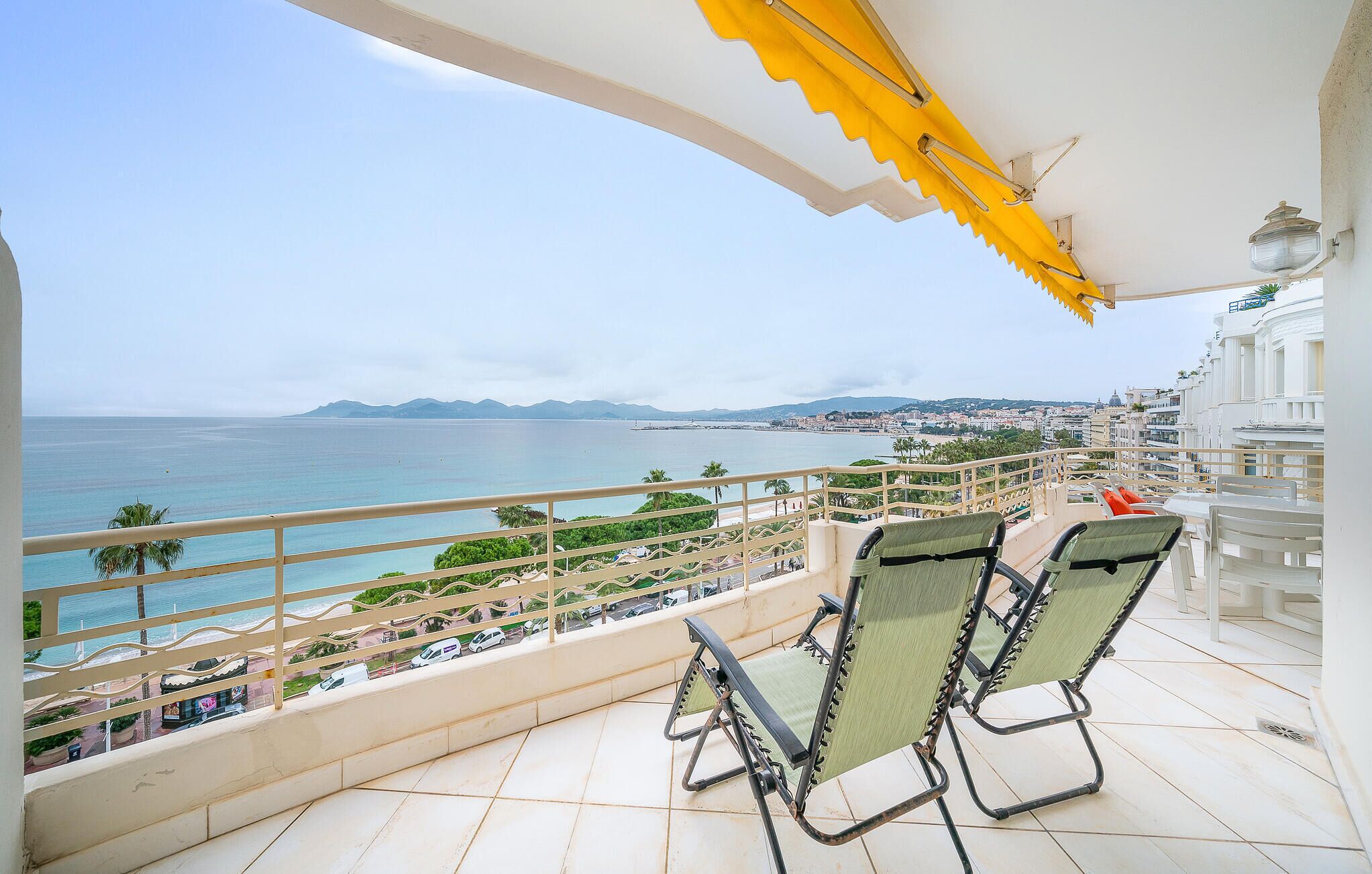 Ein Balkon mit zwei grünen Stühlen, einem gelben Sonnensegel und Blick auf das Meer und die Stadt.