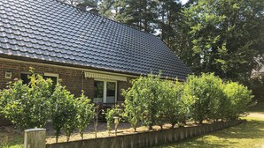 Une maison en briques avec un toit en tuiles, un porche à l’avant et un petit jardin planté de jeunes arbres.