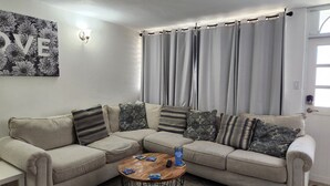 A living room with a beige sectional sofa, a wooden coffee table, and a wall-mounted light fixture. - Discover Beautiful Beaches in Fajardo! 3 Bed, 2 Bath home, Pool, BBQ, full AC! (Fajardo)