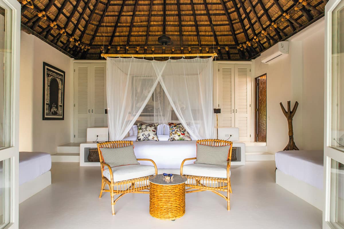 A room with a bed, two wicker chairs, a small round table, a canopy bed, and a ceiling with a thatched roof.