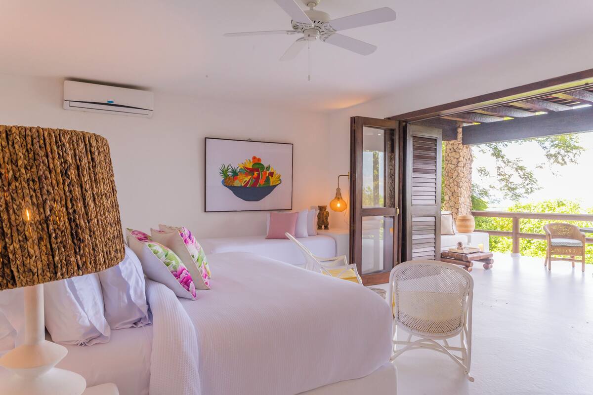 Une chambre à coucher comprenant un lit, un ventilateur de plafond, un climatiseur, un tableau et un balcon avec des meubles en osier.