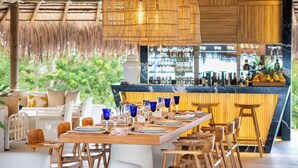 Een restaurant met een lange houten tafel gedekt voor diner, een bar met flessen en uitzicht op groen buiten.
