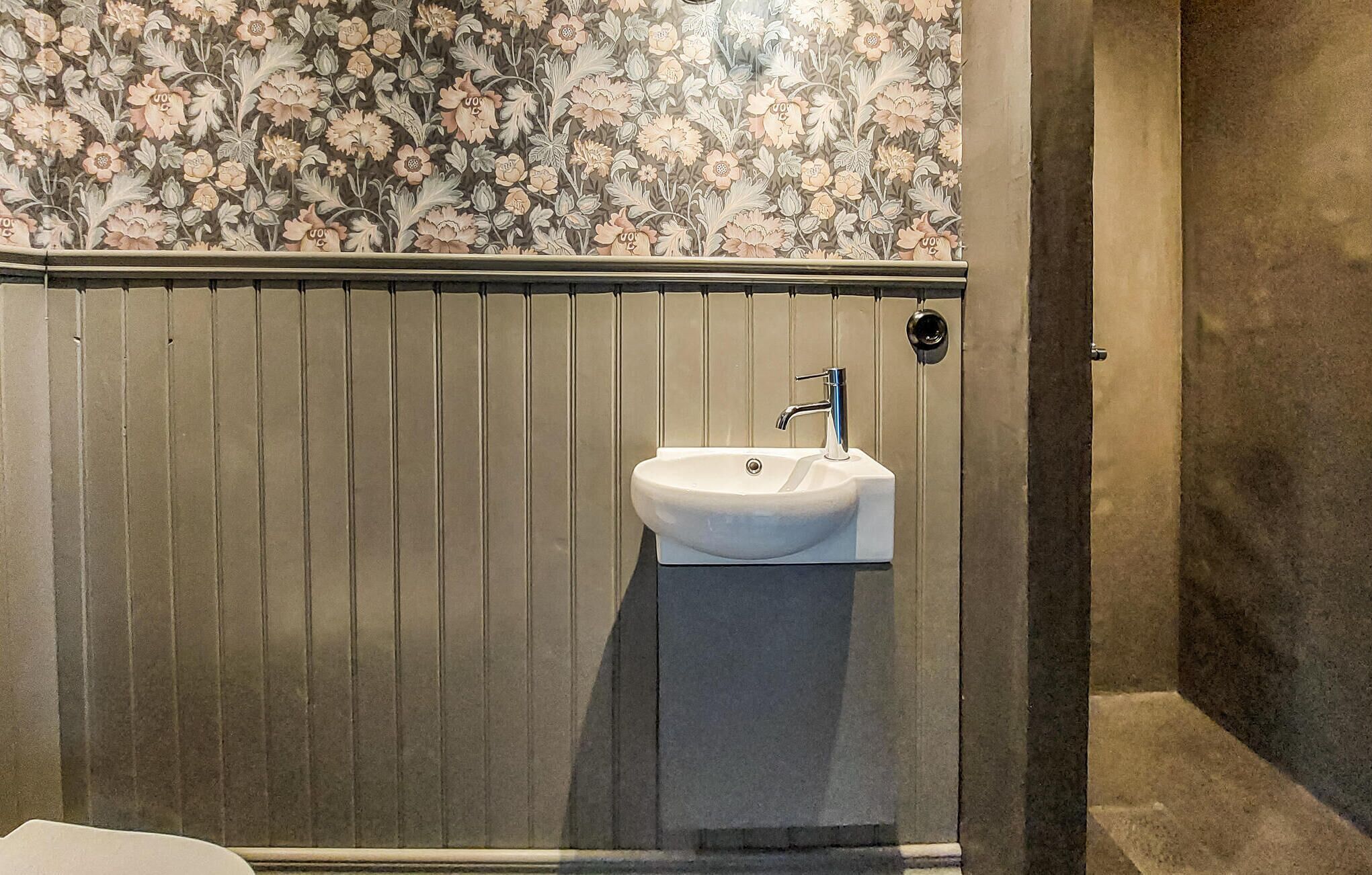 Un baño con un lavamanos blanco, diseño de papel tapiz floral y pared de paneles de madera.