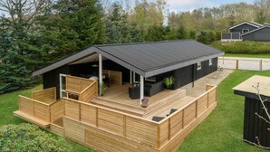 En moderne udendørs terrasse med et overdækket siddeområde, omgivet af en træterrasse og en græsplæne.