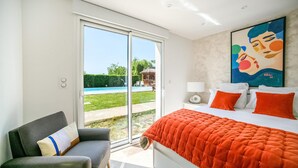 A bedroom with a bed, a chair, a painting, and a view of a swimming pool.