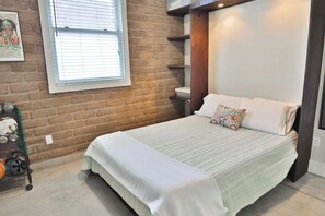 A bedroom with a brick wall, a window with blinds, a bed with a white comforter, and a shelf unit. - Barrio Viejo, Charming Historic Downtown Home (Tucson)