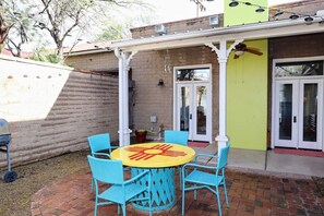 A patio with a round table and chairs, a barbecue grill, and a door leading to a building with a yellow wall. - Barrio Viejo, Charming Historic Downtown Home (Tucson)