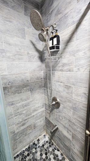 A shower with a rain showerhead and a handheld showerhead, a towel rack with bottles, and a hexagonal tile floor.