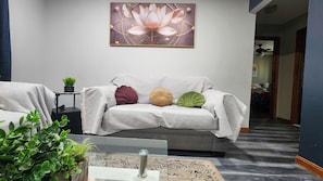 A living room with a grey sofa, a framed artwork above it, a small table with a plant, and a glass coffee table with a plant.