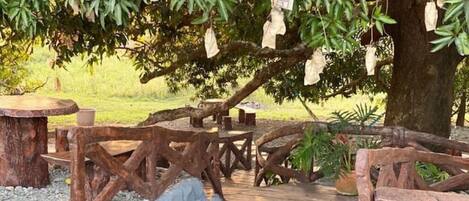 A rustic outdoor seating area with wooden tables and benches under a tree, surrounded by gravel and greenery.