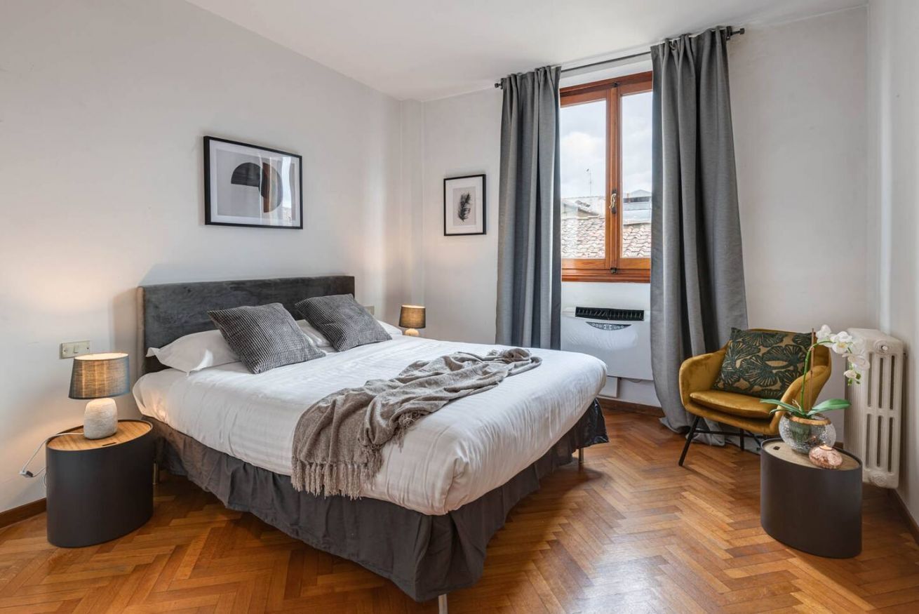 Een moderne slaapkamer met een groot bed, twee bedlampen, een stoel met een plant en een raam met gordijnen.