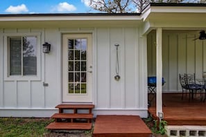 Una casa blanca con puerta principal, ventana con persianas, ducha, patio con mesa y sillas y un pequeño jardín.