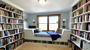 Eine gemütliche Leseecke mit integriertem Bett, umgeben von Bücherregalen voller Bücher, einem Fenster mit Aussicht und einer Deckenleuchte.