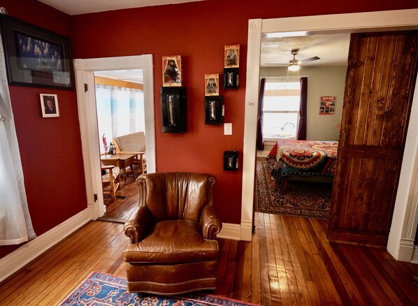 Une pièce avec un fauteuil en cuir marron, un tapis à motifs et un lit recouvert d’une couverture colorée. Des tableaux encadrés ornent le mur, et une porte donne accès à une autre pièce.