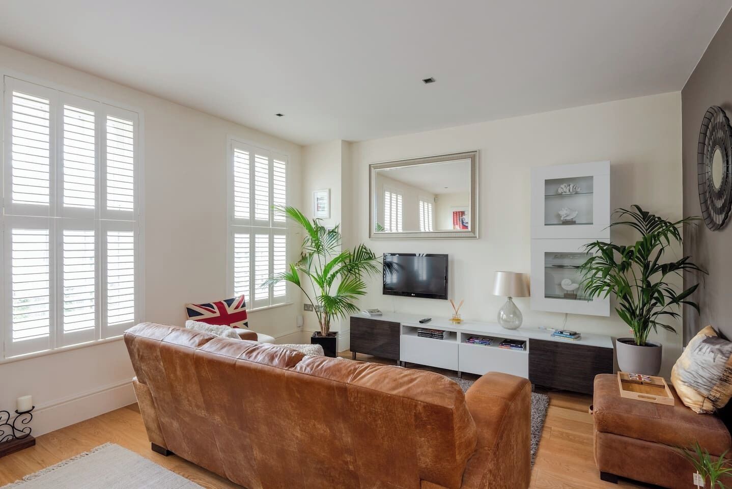 A living room with a brown sofa, a smaller armchair, a TV, a mirror, and two potted plants.