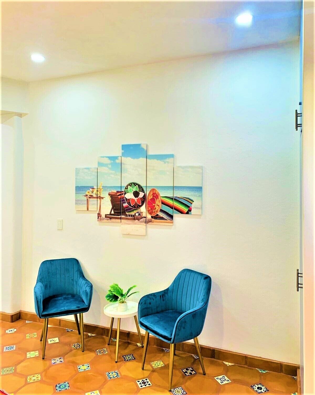 A room with two blue chairs, a small table with a plant, and a wall-mounted artwork depicting a beach scene.