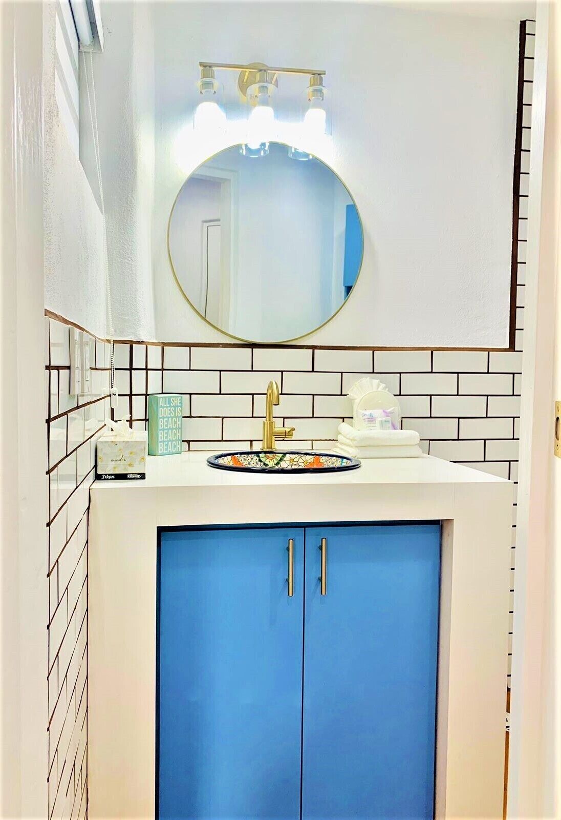 Ein Badezimmer mit einem weißen Waschbecken, einem goldenen Wasserhahn und einem blauen Schrank. Darüber befindet sich ein runder Spiegel und links daneben ein Handtuchhalter.