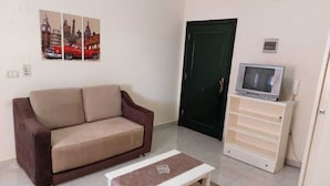 A living room with a brown sofa, a white coffee table, a television on a stand, and a door in the background. - Cozy / Comfy Studio (Hurghada)