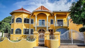 Una casa grande de dos pisos, de color amarillo, con techo de tejas rojas, balcones blancos y una entrada con reja.