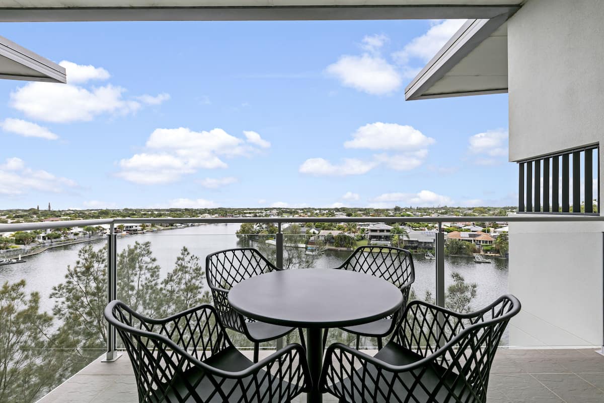 Un balcon avec une table ronde et des chaises, donnant sur une rivière où l’on aperçoit des bateaux et des bâtiments au loin.
