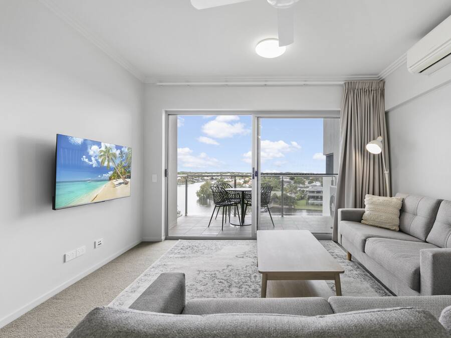 Una sala de estar moderna con un sofá gris, una mesa de centro y un televisor montado en la pared. Hay un balcón con vistas al cielo y un paisaje distante.