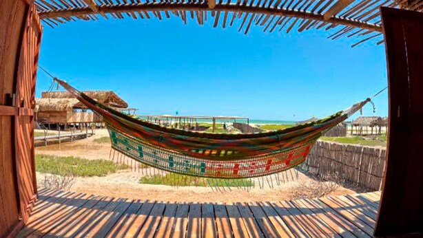 A hammock hanging between wooden beams, overlooking a beach with thatched-roof structures and a clear blue sky. - Palaa Mayapo Ecolodge (Manaure)