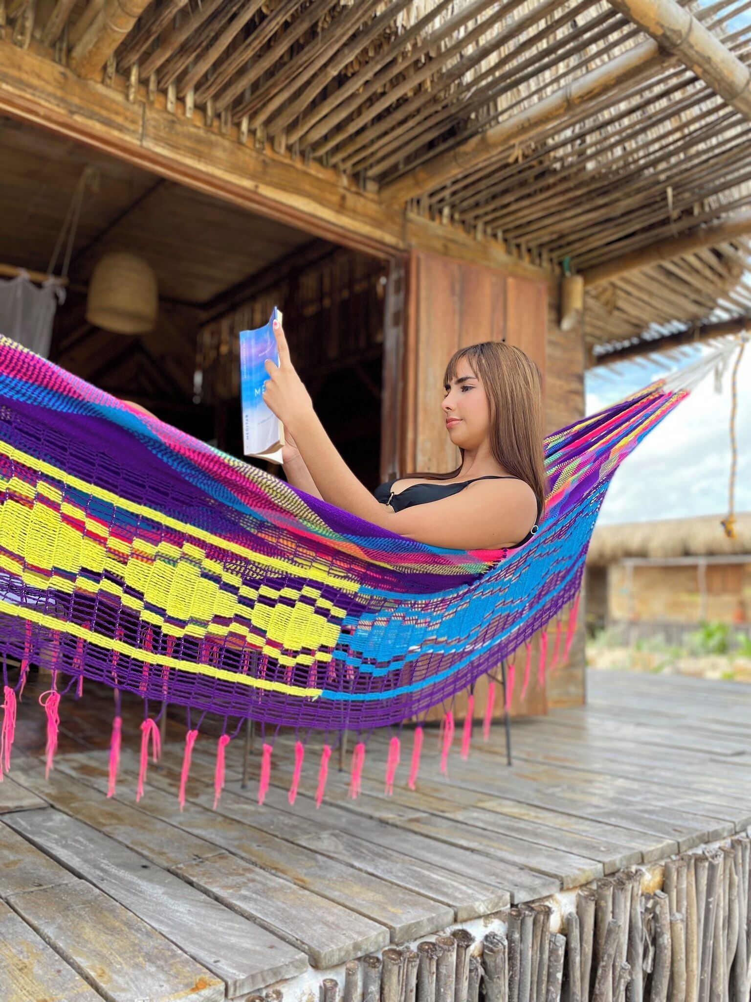 A person relaxing in a colorful hammock on a wooden deck with a thatched-roof building in the background.