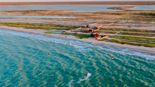 An aerial view of a coastal area with clear turquoise waters, a sandy beach, and a small settlement with buildings and boats.