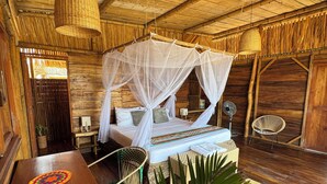A rustic wooden room with a bed under a canopy, a woven chair, a small table, and a woven floor mat.
