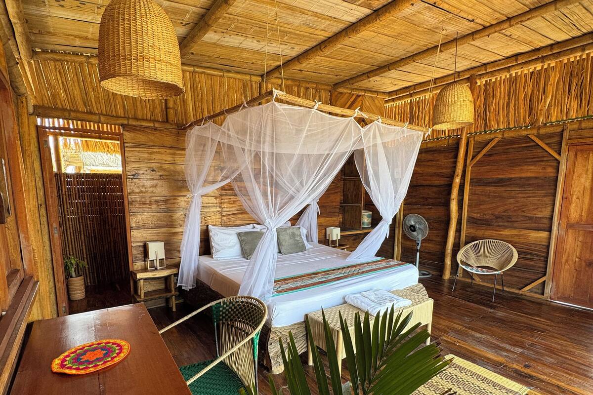 A rustic wooden room with a bed under a canopy, a woven chair, a small table, and a woven floor mat.
