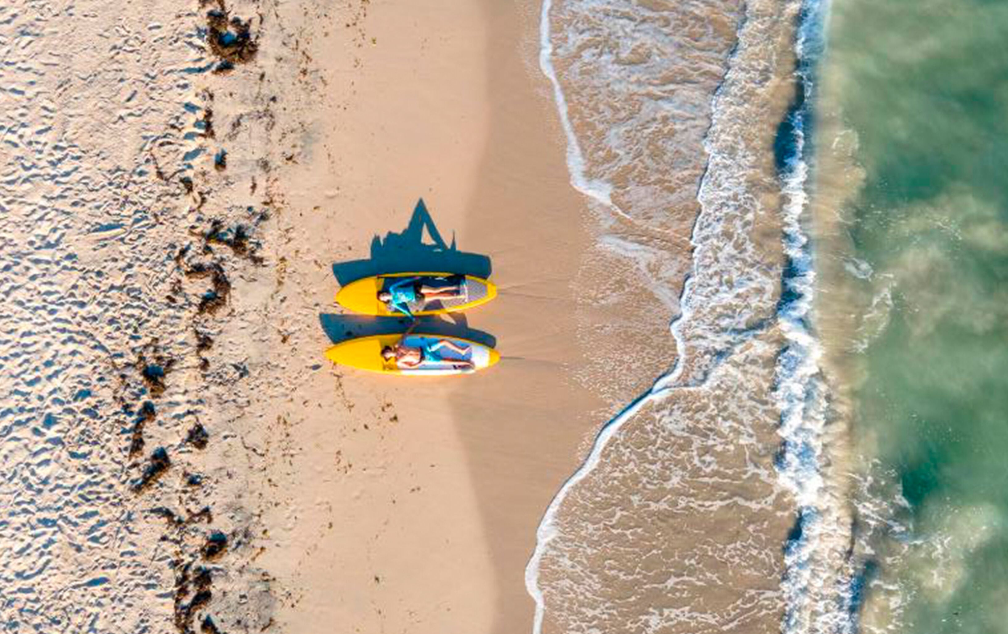 Vista aérea de uma praia com um caiaque amarelo e uma prancha de surf azul.