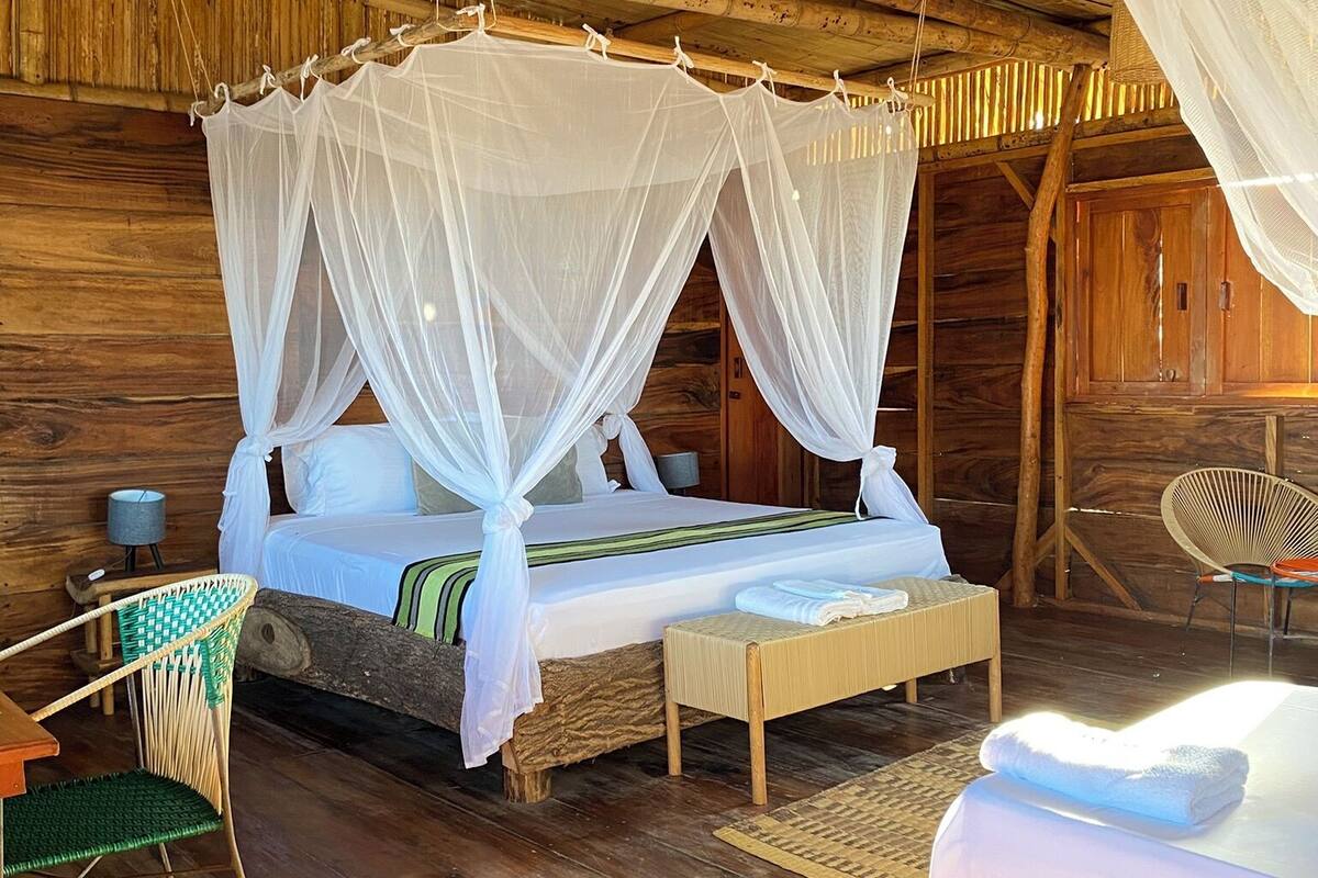 A rustic wooden room with a bed under a canopy, a woven basket hanging from the ceiling, and a chair with a woven seat.
