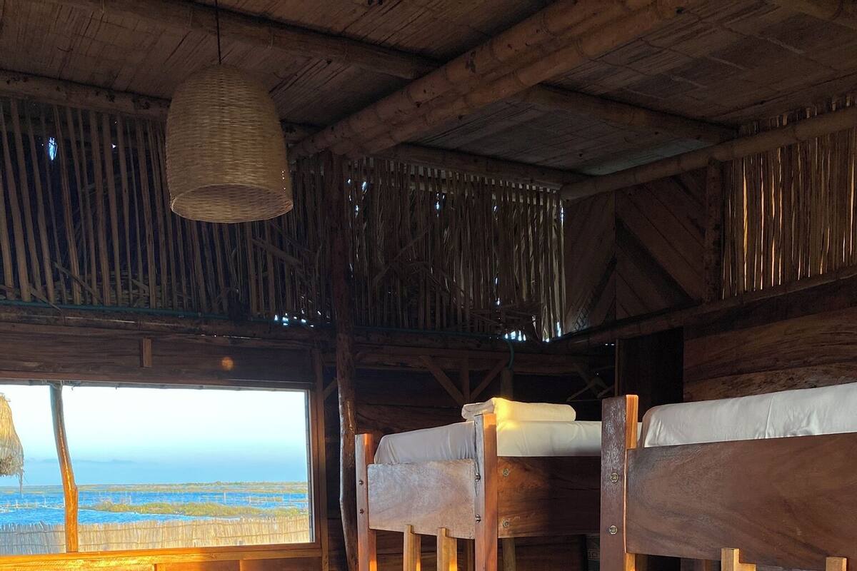 A rustic cabin with wooden bunk beds, a woven hanging light fixture, a wooden table with a chair, and a view of the beach.