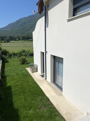 Um edifício moderno com fachada branca, antena parabólica, pequena área externa com mesa e cadeiras, e vista para um campo verde e montanhas ao fundo.