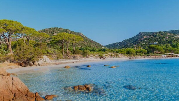 Una playa costera despejada con formaciones rocosas y colinas verdes al fondo.
