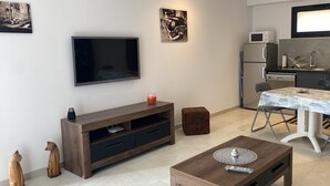 Un espacio habitable compacto con televisor, una mesa de comedor, una zona de cocina con refrigerador y microondas, y una pequeña mesa de centro.
