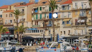 Un puerto deportivo con barcos amarrados, una iglesia con campanario y edificios con balcones y tiendas.