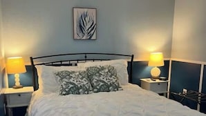 A neatly made bed with a geometric patterned bedspread, two bedside lamps, a ceiling fan, and a wall-mounted abstract art piece. - Seaside Sunsets, Our Oceanfront Home In Aripeka (Spring Hill)
