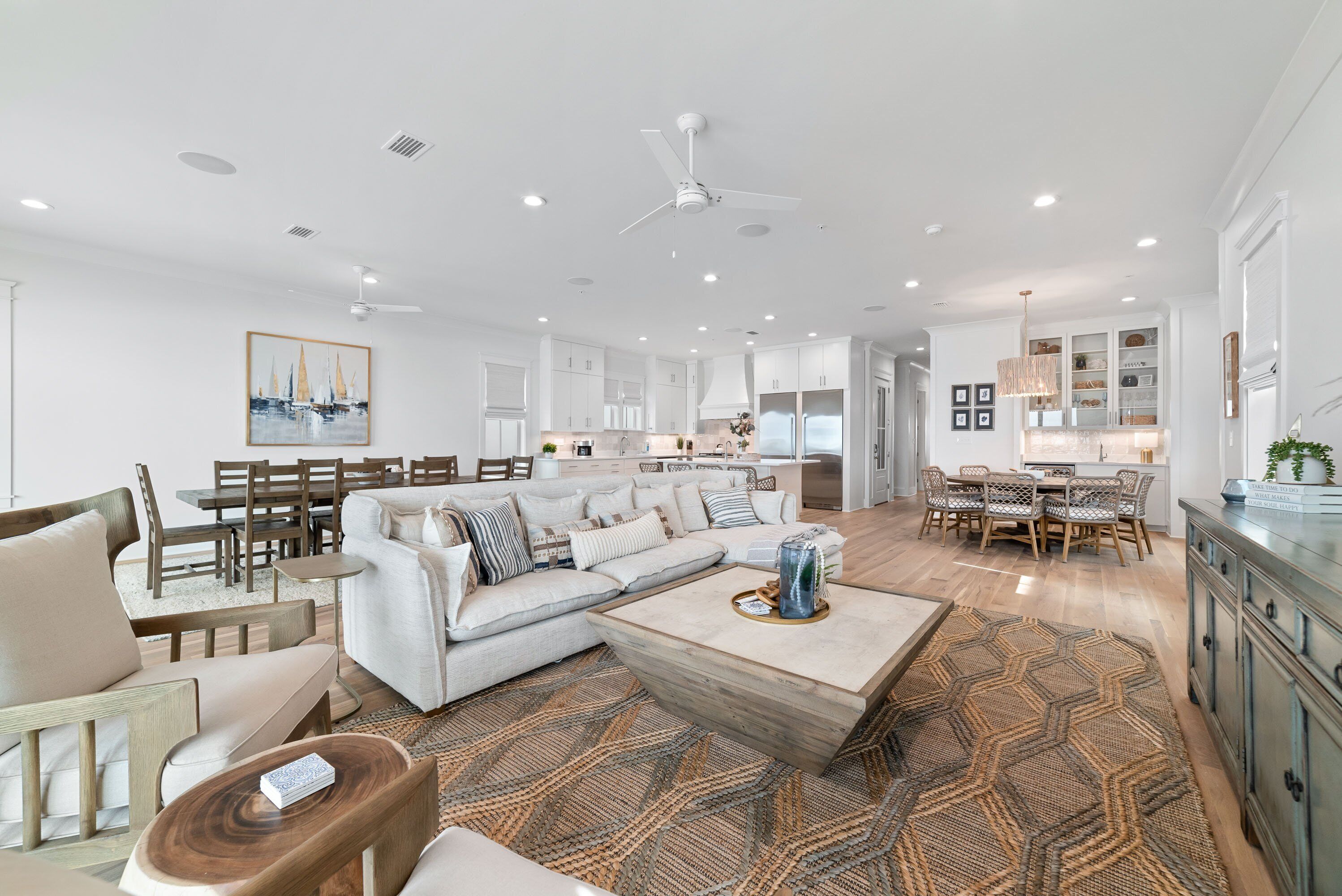A spacious living area with a large sofa, coffee table, and dining table. There is a kitchen in the background with white cabinets and a chandelier.