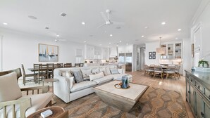 A spacious living area with a large sofa, coffee table, and dining table. There is a kitchen in the background with white cabinets and a chandelier. - BEACHFRONT, LUXURY HOUSE, PRIVATE POOL! (Orange Beach)